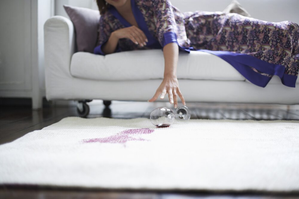 Woman picking up a wine glass after spilling red wine on the rug. 